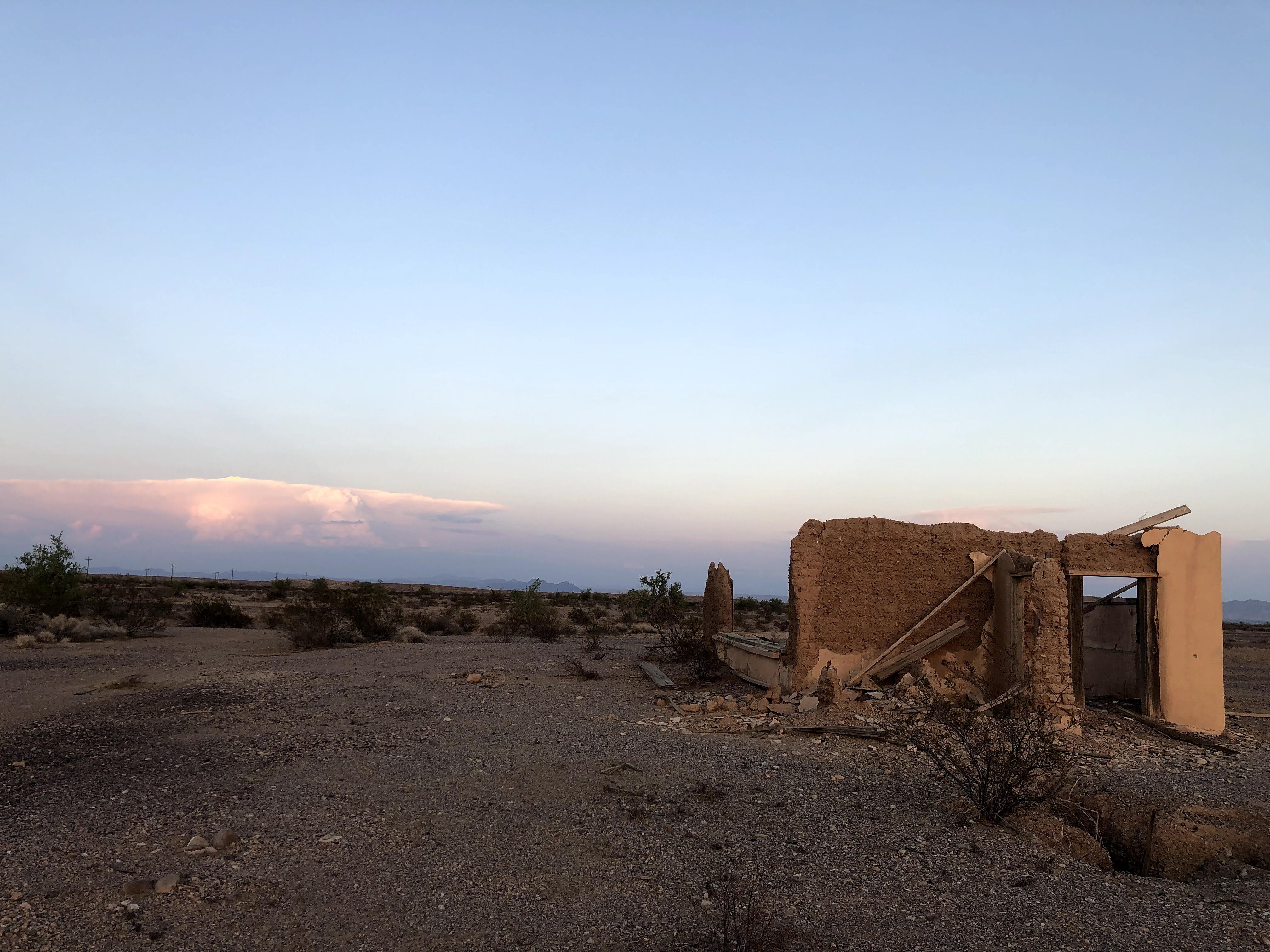 The collapsing adobe walls of an abandoned building on the open desert floor near Vidal, California, at dusk, with distant mountains and pink-lit clouds on the horizon.