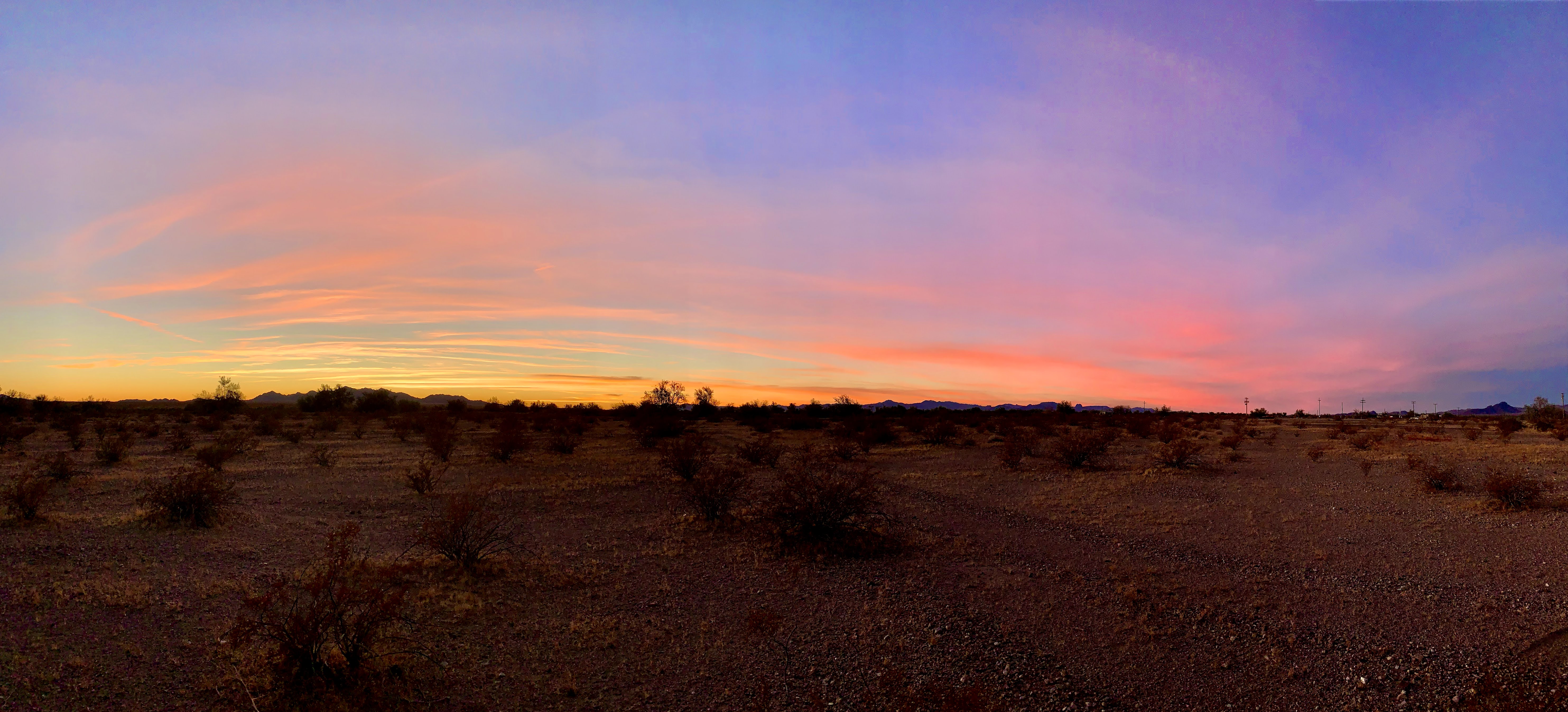 A wide panoramic sunset over the desert near Vidal, California — bands of pink, orange, and violet across the sky above silhouetted creosote and distant mountains.
